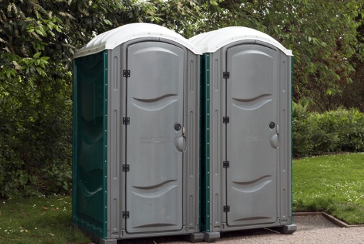 Standard gray and blue portable toilet with vent stack and secure door, standing on a construction site.