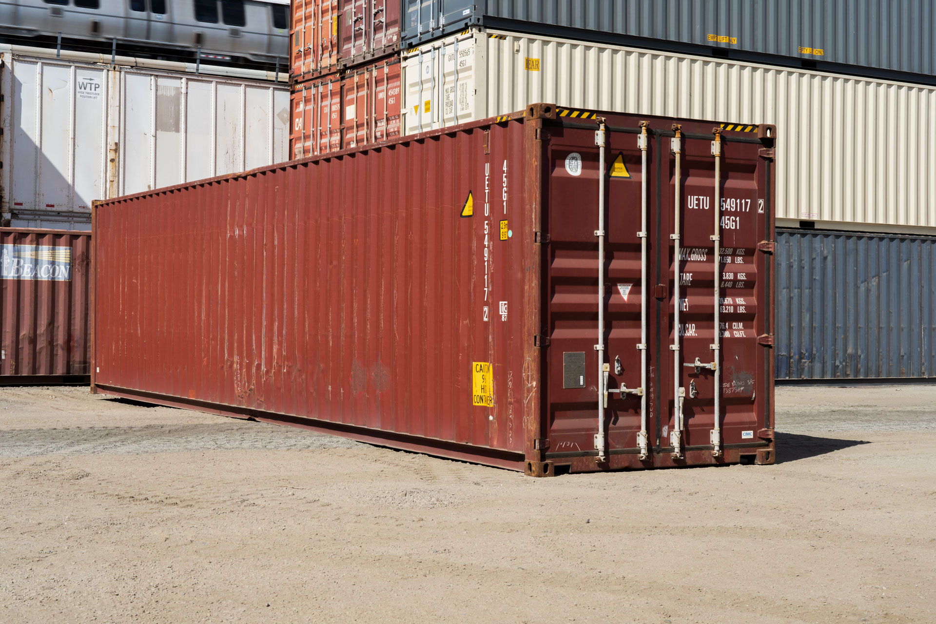 Stacked shipping containers in various colors, including red and blue, arranged in rows at an industrial storage yard under a partly cloudy sky.